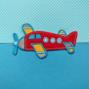 Op de afbeelding: Een rode vliegtuigapplicatie met blauwe en gele details. Het vliegtuig heeft drie ronde ramen en een propeller.
