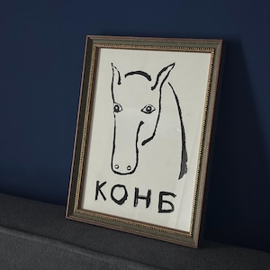 Könnte beinhalten: Ein gerahmter Kunstdruck mit einer schwarzen Tuschezeichnung eines Pferdekopfes auf weißem Hintergrund. Unter dem Pferd ist der kyrillische Text "КОНБ" in fetten schwarzen Buchstaben gedruckt. Das Kunstwerk befindet sich in einem rechteckigen Rahmen mit einem strukturierten braunen Rand und einem dünnen goldenen Innenbesatz.