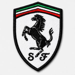Può includere: Una toppa ricamata a forma di scudo con sfondo bianco e la sagoma nera di un cavallo rampante al centro. Il bordo superiore presenta strisce orizzontali verde, bianca e rossa. Le lettere S e F sono ricamate in nero nella parte inferiore.
