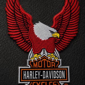 May include: An embroidered patch featuring a red eagle with white head and yellow beak perched atop a black and orange Harley-Davidson Motor Cycles logo. The emblem includes the text MOTOR, HARLEY-DAVIDSON, and CYCLES with a registered trademark symbol.