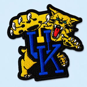 May include: An embroidered patch featuring a stylized yellow wildcat with a red tongue and white teeth, positioned above and behind the blue block letters UK. The design is outlined in black thread, creating a bold, high-contrast graphic emblem.