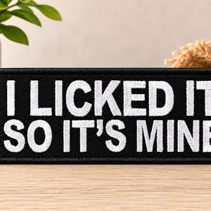 Może przedstawiać: Prostokątna, czarna haftowana naszywka z obszytymi brzegami, zawierająca biały napis I LICKED IT SO IT'S MINE wykonany pogrubioną czcionką bezszeryfową. Naszywka ma płaską, przypominającą tkaninę fakturę i jest zorientowana poziomo.