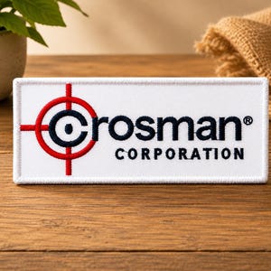 May include: A rectangular white embroidered patch featuring the Crosman Corporation logo. The design includes a red and black circular target graphic to the left of the black embroidered text that reads Crosman Corporation.
