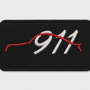 May include: A rectangular black embroidered patch featuring a white stitched number 911. A continuous red stitched line forms the silhouette of a sports car profile that overlays the white numbers, creating a distinct graphic design on the fabric.