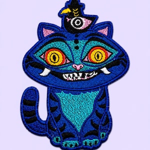 Puede incluir: Un parche bordado que muestra un gato azul con grandes ojos amarillos, una amplia sonrisa con dientes blancos visibles y rayas negras. Un pequeño pájaro negro con un solo ojo se posa sobre la cabeza del gato. El diseño utiliza hilo azul, turquesa, amarillo y rosa.