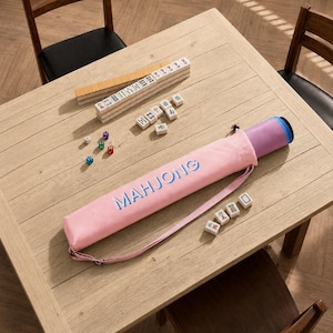 May include: A light pink cylindrical carrying case with the word MAHJONG printed in blue, containing a rolled mat. Scattered on the surface are white mahjong tiles with black and green characters, and four small colorful dice in red, green, and blue.