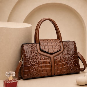 May include: A brown handbag with a glossy crocodile-embossed texture, featuring a structured top handle, a flap closure with a central vertical panel, and side attachments for a shoulder strap. The bag has a polished finish and dark brown trim.