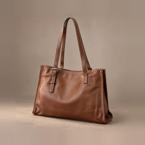 May include: A medium brown leather tote bag featuring two long shoulder straps, a rectangular silhouette, and a decorative vertical leather strap detail on the front panel secured with a small silver-toned metal buckle and matching side rivets.