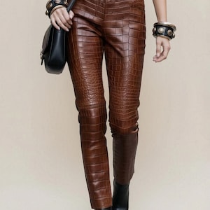 May include: Brown trousers featuring a textured crocodile-skin pattern, a matching brown belt with a rectangular buckle, a white t-shirt, a black shoulder bag, black pointed-toe ankle boots, and two wide black bracelets with metallic studs.