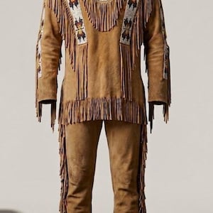 May include: A tan suede outfit consisting of a long-sleeved pullover shirt and matching trousers. The shirt features long fringe along the sleeves and hem, with white beaded panels displaying geometric patterns in black, orange, and yellow.