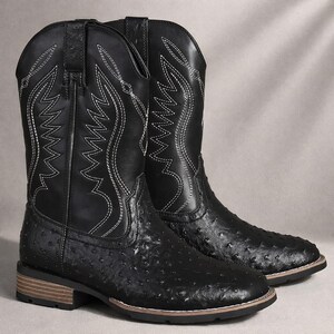 May include: A pair of black cowboy boots featuring a textured, quill-like pattern on the foot and a smooth shaft with white decorative stitching. The boots have a square toe, a low stacked heel, and pull tabs at the top of the shaft.