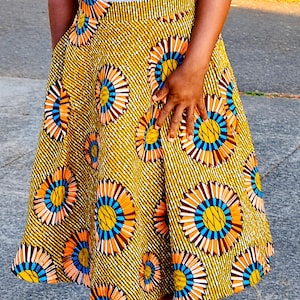 May include: A high-waisted midi skirt featuring a vibrant mustard yellow background with a repeating pattern of circular sunburst motifs in shades of orange, blue, and white, accented by thin diagonal white stripes across the entire fabric surface.