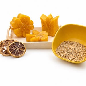 May include: Three yellow flower-shaped soap bars with visible textured particles, arranged on a small wooden tray. Beside the tray are two dried citrus slices and a yellow bowl filled with a coarse, light brown granular exfoliating substance.