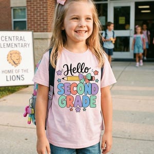 Può includere: Una t-shirt a maniche corte rosa chiaro con una stampa grafica che riporta la scritta Hello SECOND GRADE in lettere nere, viola, blu e verdi. Il design include una matita gialla, un fiocco colorato e piccole icone a forma di fiori e stelle.