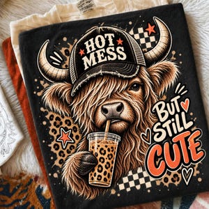 May include: A black t-shirt featuring a graphic of a Highland cow wearing a black trucker hat with the text "HOT MESS". The cow is drinking from a leopard-print cup. The design includes leopard spots, stars, and the text "BUT STILL CUTE".
