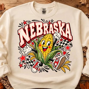 May include: A cream-colored crewneck sweatshirt featuring a graphic of a cartoon corn cob character with arms and legs, wearing red sneakers. The text NEBRASKA is printed in large red letters above the corn, surrounded by stars and checkered flags.