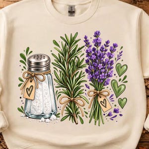 Puede incluir: Una sudadera de cuello redondo color crema con un estampado gráfico de un salero de cristal, una rama de romero verde y un manojo de lavanda morada. Cada elemento está atado con cuerda y una pequeña etiqueta con un corazón negro dibujado.