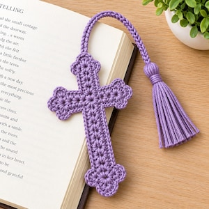 Puede incluir: Un marcapáginas de ganchillo color lavanda con forma de cruz y borde festoneado decorativo. El marcapáginas cuenta con un cordón trenzado unido a una borla color lavanda a juego, sobre las páginas abiertas de un libro con texto.