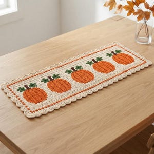 May include: A rectangular crochet table runner in cream yarn featuring five orange pumpkins with green stems and leaves. The runner has a scalloped edge border and is crafted with a textured stitch pattern, displaying a repeating pumpkin motif.