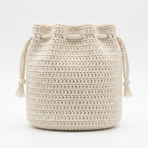 Può includere: Una borsa a secchiello all'uncinetto color crema caratterizzata da un motivo a maglia denso e strutturato. La borsa ha una forma cilindrica con chiusura a coulisse sulla parte superiore, rifinita con due cordini intrecciati che pendono ai lati con nappe.