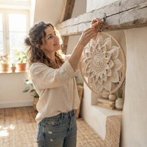 May include: A cream-colored crochet wall hanging featuring a layered, petal-like mandala design secured within a circular wooden hoop. The intricate fiber art piece displays a central circular pattern surrounded by multiple pointed, overlapping layers.