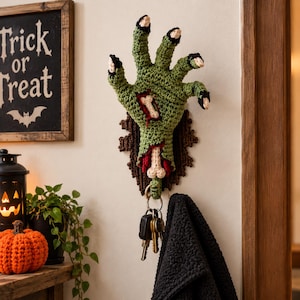 Puede incluir: Un gancho de pared con forma de mano de zombi tejida en color verde, con un detalle de hueso expuesto, uñas negras y una base texturizada de color marrón oscuro. La mano incluye una anilla metálica en la muñeca que sostiene un juego de llaves.