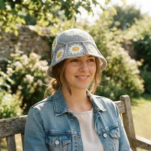 May include: A light gray crochet bucket hat featuring a pattern of white and yellow daisy motifs. The hat has a wide, downward-sloping brim and a small, circular wooden tag attached to the front side, showcasing a detailed handmade textile design.