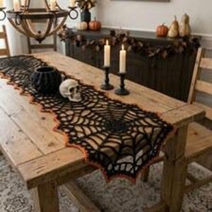 Peut inclure: Un chemin de table en crochet noir avec un motif de toile d'araignée et une bordure orange. Le chemin est posé sur une table en bois et décoré d'un petit crâne blanc et d'un bol noir texturé, créant une décoration sombre et saisonnière.