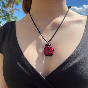 May include: A necklace featuring a ladybug pendant with a bright red body and black spots, suspended from a simple black cord. The ladybug has a glossy finish and a small black head, hanging centered against a plain black fabric garment.
