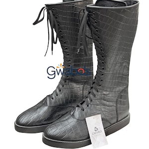 May include: A pair of black knee-high boots featuring a textured crocodile skin pattern, black laces, and a flat sole with white stitching. A white rectangular tag with the text GWABO CLOSET is attached to the front of the right boot.