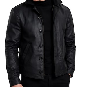 May include: A black jacket featuring a pointed collar, long sleeves, and a front button closure. The jacket is worn over a black mock neck shirt. The material has a smooth, matte finish with visible seams along the chest and side pockets.