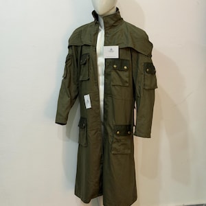 May include: An olive green long coat featuring a high collar, multiple utility pockets with dark brown flaps and gold-toned buttons, and a cape-style overlay across the shoulders. The garment includes two white rectangular tags attached to the front.