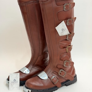 May include: A pair of knee-high brown boots featuring multiple side buckles, a silver-toned metal toe cap, and a thick black lug sole. A white tag with the text GWABO CLOTHES is attached to the boots, which also feature vertical stitching details.