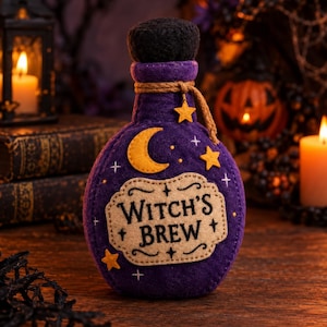 Puede incluir: Una decoración de fieltro morado con forma de botella y tapón de fieltro negro. La parte frontal tiene una etiqueta beige con el texto "WITCH'S BREW" en negro, decorada con una luna creciente de fieltro amarillo, estrellas y cordel marrón.