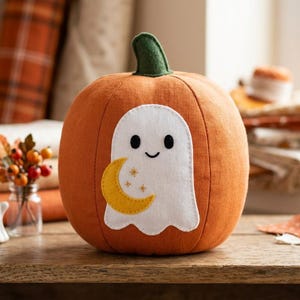 May include: A round orange fabric pumpkin with a dark green stem. The front features a white ghost appliqué holding a yellow crescent moon with three small stars. The ghost has two black oval eyes and a simple curved line for a mouth.
