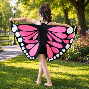 May include: Butterfly wings costume accessory featuring a black base with vibrant pink wing panels and white circular accents along the outer edges. The fabric wings are attached to a central black harness worn over the shoulders with thin straps.