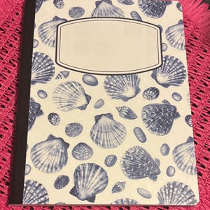 May include: A rectangular notebook with a white cover featuring a repeating pattern of blue seashells and small stones. The notebook has a dark brown spine and a large, blank, light-coloured rectangular label with a thin border centred on the front.