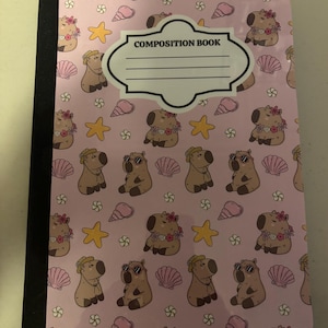 May include: A composition book with a light pink cover featuring a repeating pattern of brown capybaras, yellow starfish, pink seashells, and small white flowers. A white label with a black border and the text COMPOSITION BOOK is centered.