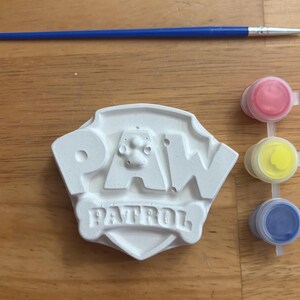 May include: A white plaster Paw Patrol shield-shaped figure, a blue paintbrush, and a strip of three small paint pots containing red, yellow, and blue paint. The Paw Patrol logo is embossed on the front of the white plaster figure.