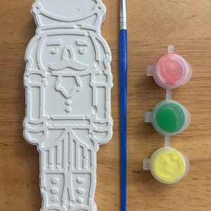 May include: A white plaster nutcracker figurine with embossed details, a blue paintbrush with orange bristles, and a strip of three small plastic paint pots containing red, green, and yellow paint for a craft project.