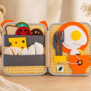 May include: A felt play set featuring a folding book design with food items including a slice of bread, tomato, cheese, and dumplings on one side, and a plate with a fried egg, a fork, a knife, and an orange teacup on the other side.