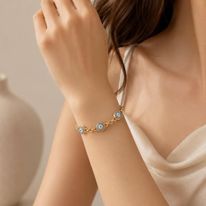 May include: A gold chain bracelet featuring multiple circular charms, each designed with a central dark blue dot surrounded by concentric rings of light blue and white, creating a repeating eye-like pattern along the length of the metal link chain.