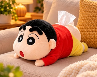 May include: A plush tissue box cover shaped like the character Shin-chan, featuring a large head with black hair, thick eyebrows, and a red shirt with yellow shorts. A white tissue is pulled from the top of the plush, which is lying flat.
