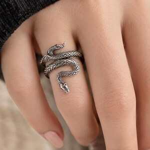 May include: A silver-toned ring shaped like a coiled snake with detailed scale textures wrapping around the finger. The metal has an oxidized finish in the crevices to highlight the serpent's form, with the head resting near the knuckle.