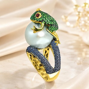 May include: A gold ring featuring a large, round, iridescent white pearl held by a frog figure encrusted with green crystals and a single red crystal eye. The band is split and paved with small, dark blue crystals, creating a textured appearance.