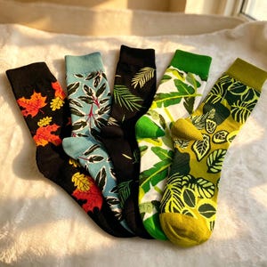 May include: Five pairs of crew socks featuring various botanical and seasonal patterns. The designs include black socks with orange and yellow maple leaves, light blue socks with dark leaves, black socks with green ferns, white socks with green palm leaves, and olive green socks with dark leaf prints.