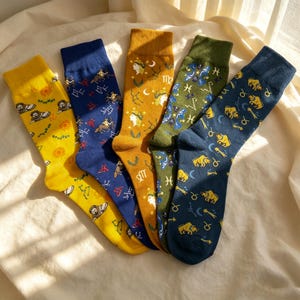 May include: Five pairs of crew socks featuring zodiac-themed patterns. The socks are yellow with lion motifs, blue with archer figures, mustard with maiden illustrations, olive green with fish symbols, and dark blue with bull graphics.