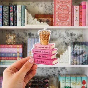 May include: A die-cut sticker featuring a stack of four pink books topped with an iced coffee in a clear plastic cup. The book spines are labeled with the text only one bed, enemies to lovers, fake dating, and slow burn in dark pink font.
