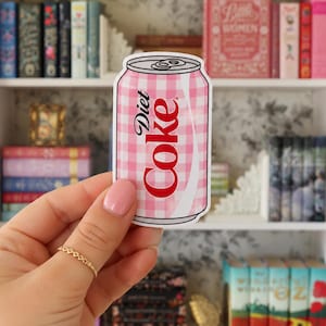May include: A die-cut sticker shaped like a soda can featuring a pink and white gingham pattern. The text Diet Coke is printed in red vertically along the side of the can. The sticker has a white border and a silver-colored top with a pull tab.