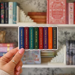 May include: A rectangular sticker featuring a row of eight colorful book spines with gold lettering. The titles include Throne of Glass, Crown of Midnight, The Assassin's Blade, Heir of Fire, Queen of Shadows, Empire of Storms, Tower of Dawn, and Kingdom of Ash.